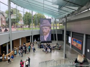 Bezoekers in de entreehal van het Van Gogh Museum Amsterdam, met een grote digitale weergave van Van Goghs zelfportret op de voorgrond en een kleurrijk schilderij zichtbaar op de achtergrond