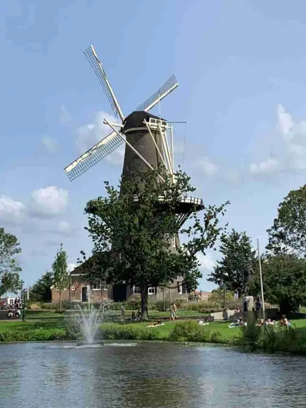 Dit is een afbeelding van windmolen De Vlieger, gelegen in een levendig park in het centrum van Leiden, met mensen die ontspannen bij een fontein op een zonnige dag.