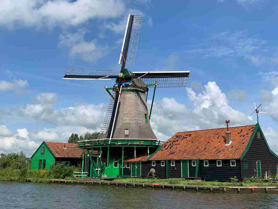 Traditionele windmolen in Zaanse Schans met groen houten gebouw, gelegen aan de waterkant onder een blauwe lucht met wolken.