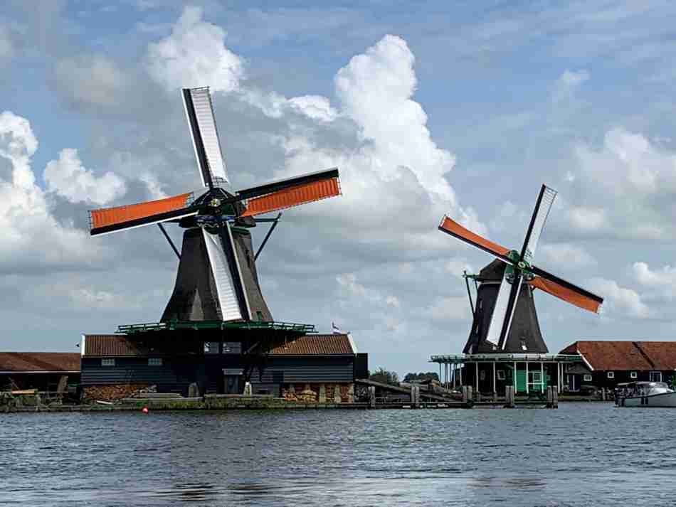Twee karakteristieke Nederlandse windmolens met oranje wieken in Zaanse Schans, weerspiegeld in het kalme water, onder een dynamische wolkenlucht.